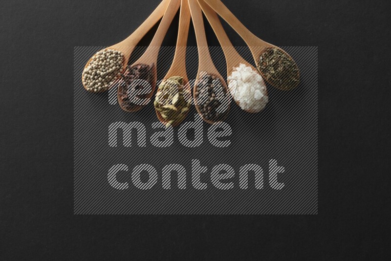 wooden spoons filled with white peppers, cloves, cardamom, salt, black peppers and basil on black flooring