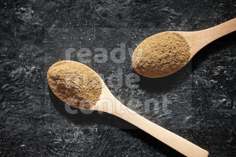 Wooden spoons full of allspice powder on a textured black flooring