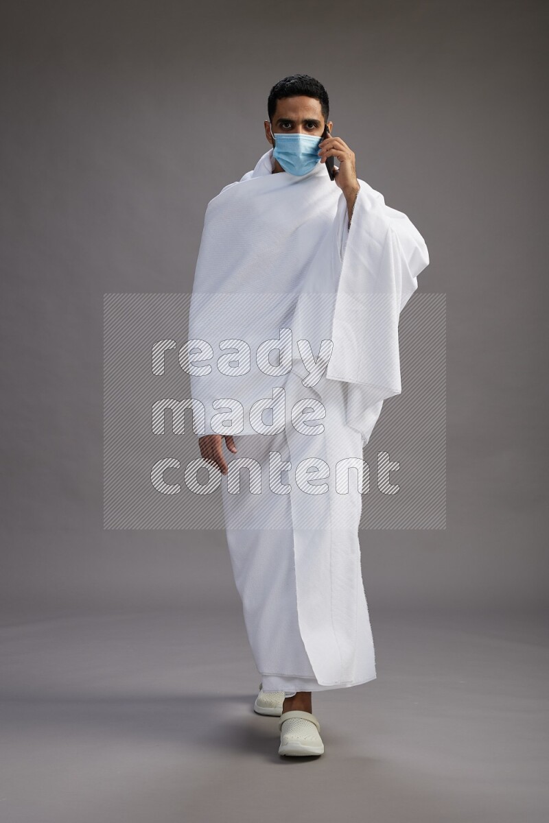A man wearing Ehram with face mask Standing talking on phone on gray background