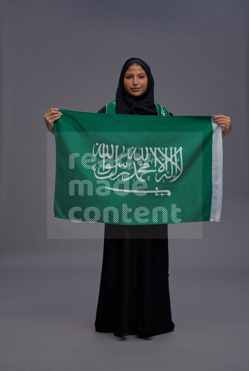 Saudi woman wearing Abaya standing holding Saudi flag on gray background