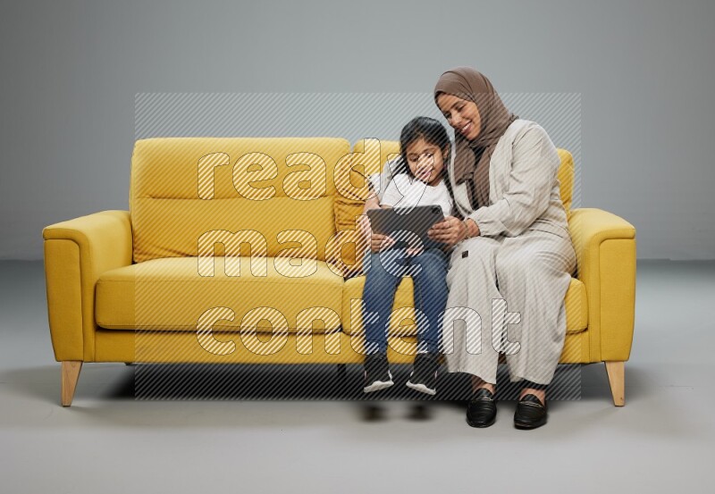 A girl with her mother on yellow sofa and watching on iPad on gray background