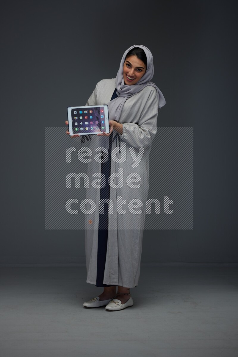 A Saudi woman wearing a light gray Abaya and head scarf standing and showing the phone's screen eye level on a grey background