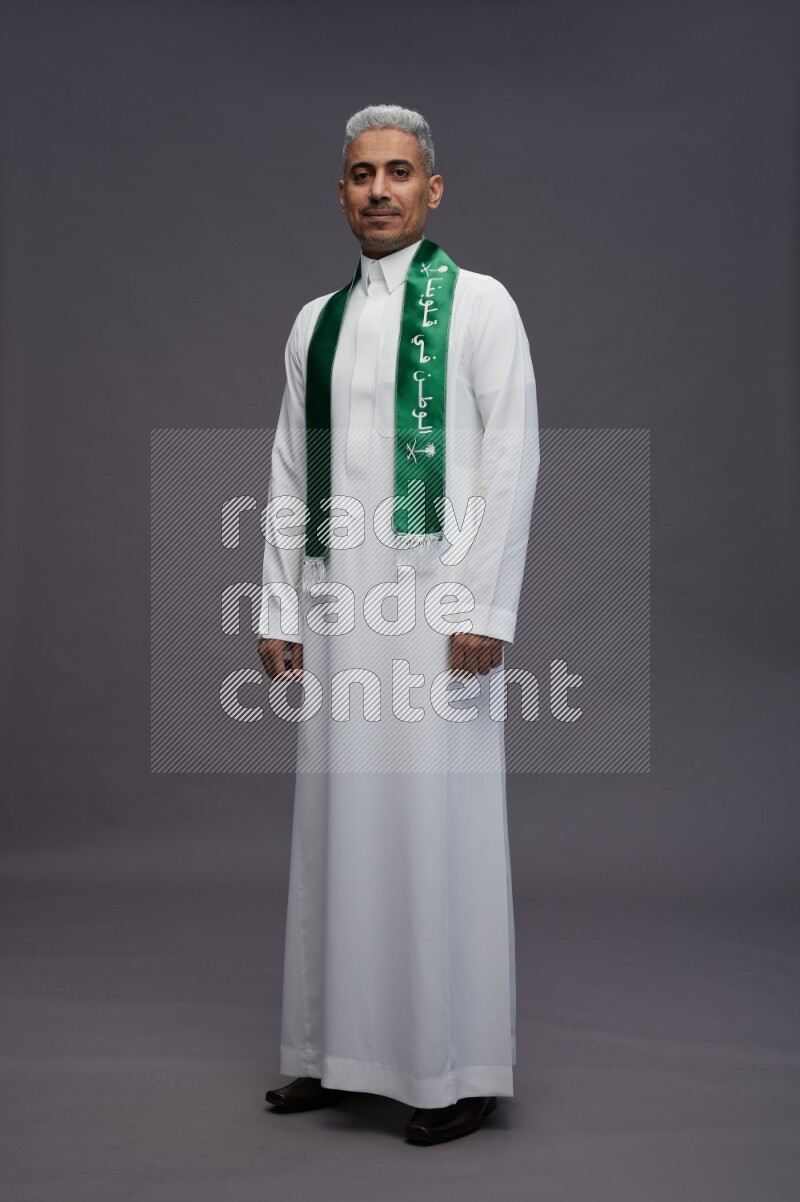 Saudi man wearing thob standing holding Saudi flag on gray background