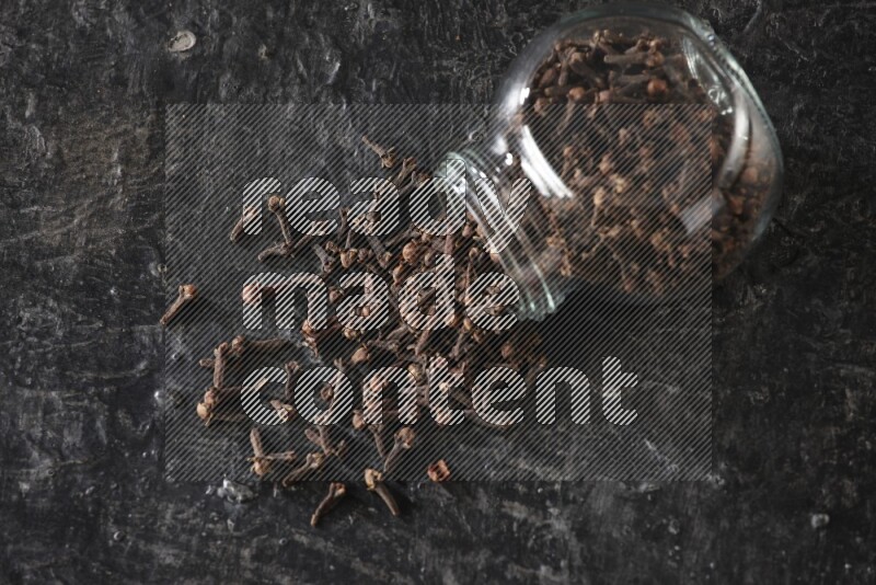 A flipped glass spice jar full of cloves and cloves came out of it on textured black flooring