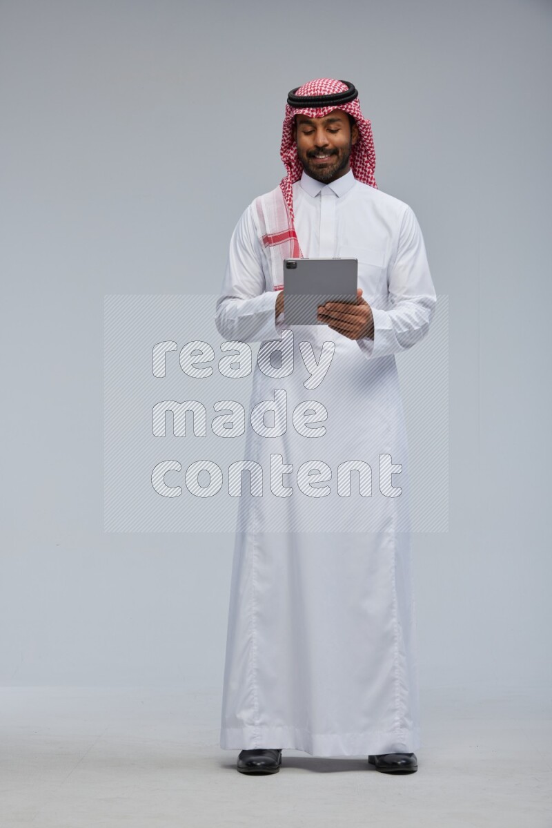 Saudi man Wearing Thob and shomag standing working on tablet on Gray background