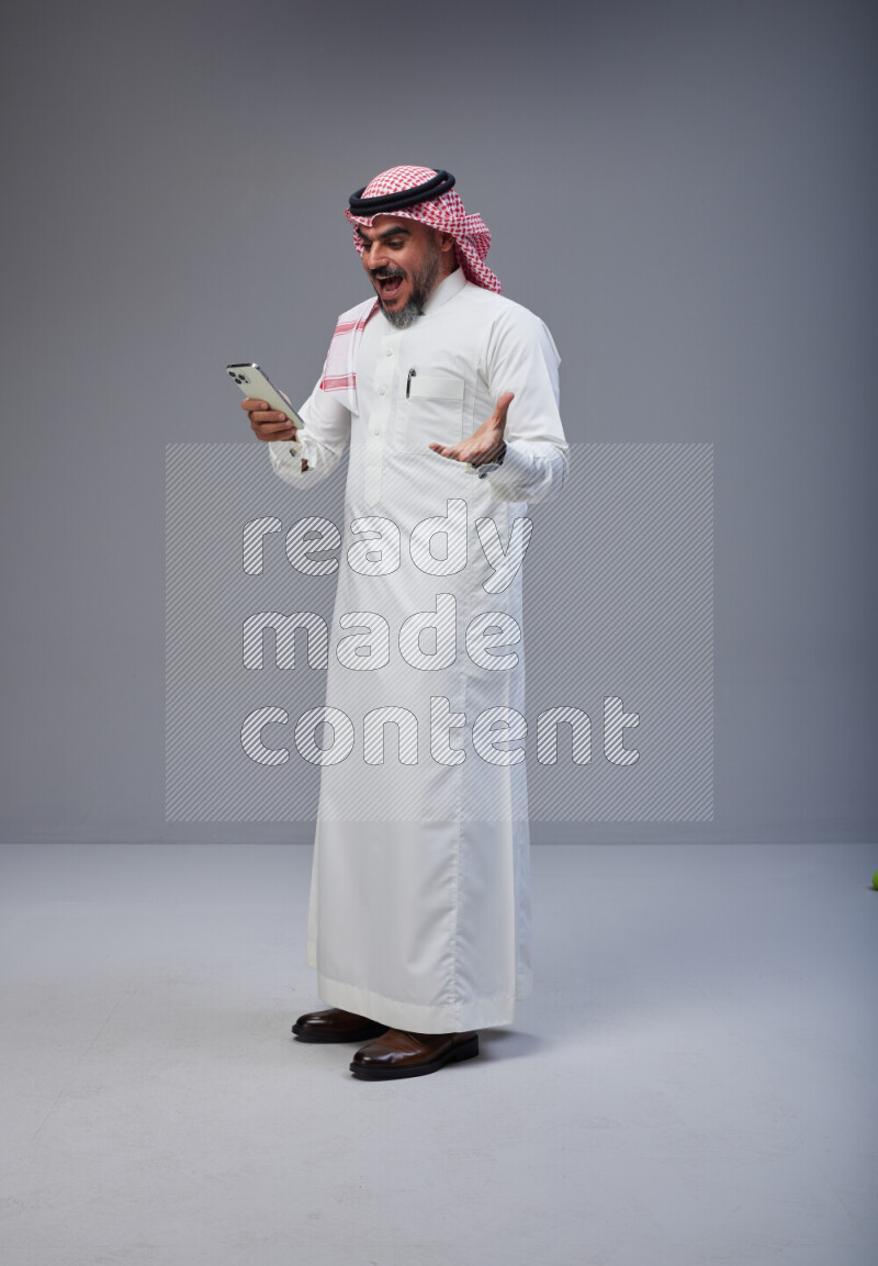 Saudi man Wearing Thob and red Shomag standing texting on phone on Gray background