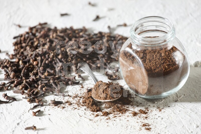 A glass spice jar and a metal spoon full of cloves powder and cloves spread on textured white flooring