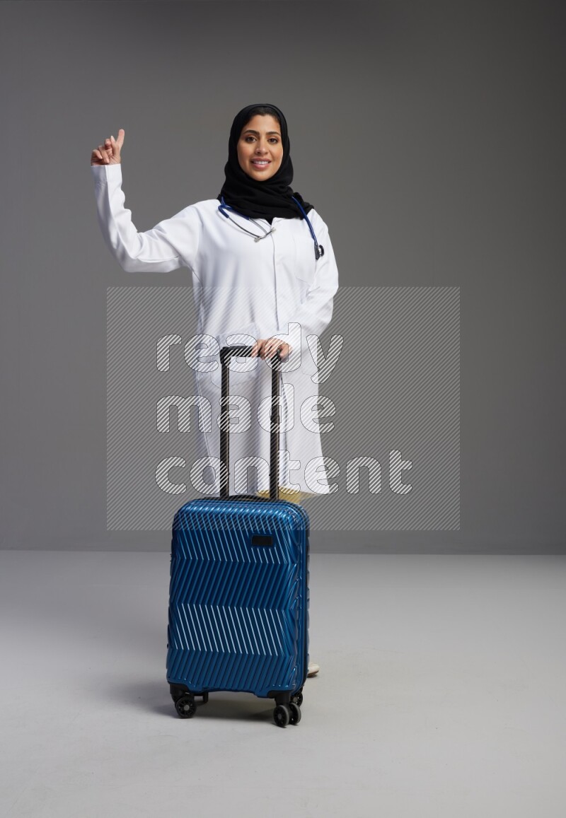 Saudi woman wearing lab coat with stethoscope standing holding Travel bag on Gray background