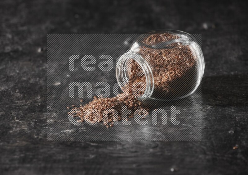 A glass spice jar full of flaxseeds flipped and seeds spread out on a textured black flooring