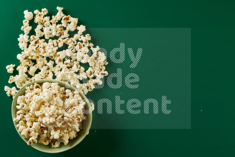 A green ceramic bowl full of popcorn with popcorn beside it on a green background in a top view shot