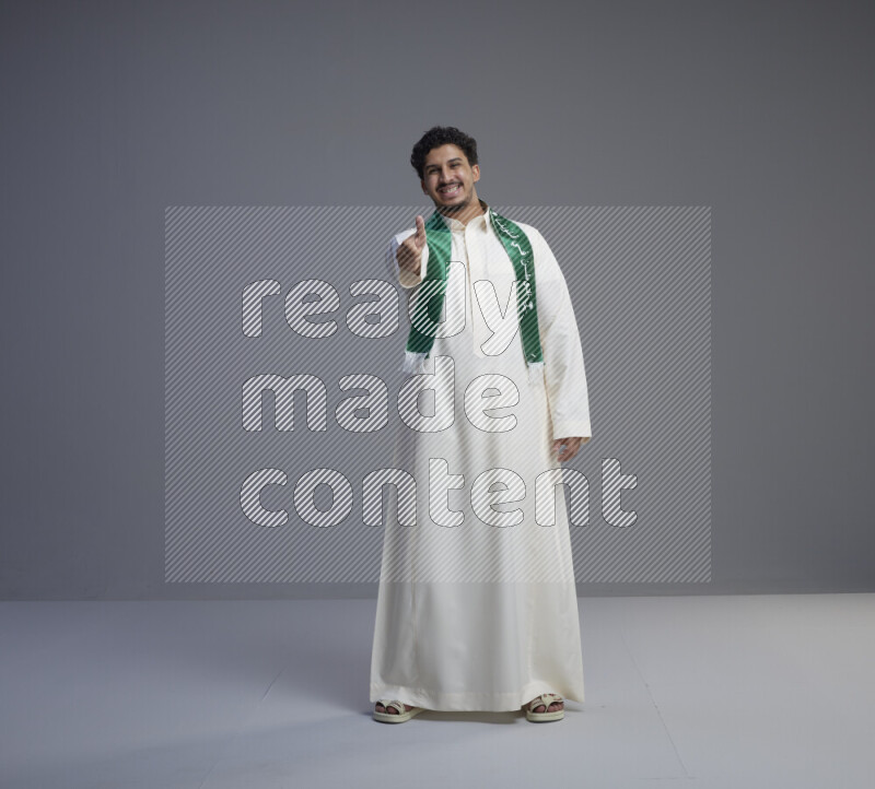 A Saudi man standing wearing thob and Saudi flag scarf on gray background
