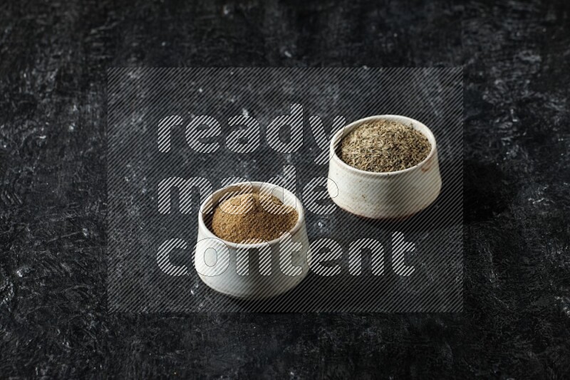 2 beige bowls, one full of cumin powder and the other full of seeds on a textured black flooring