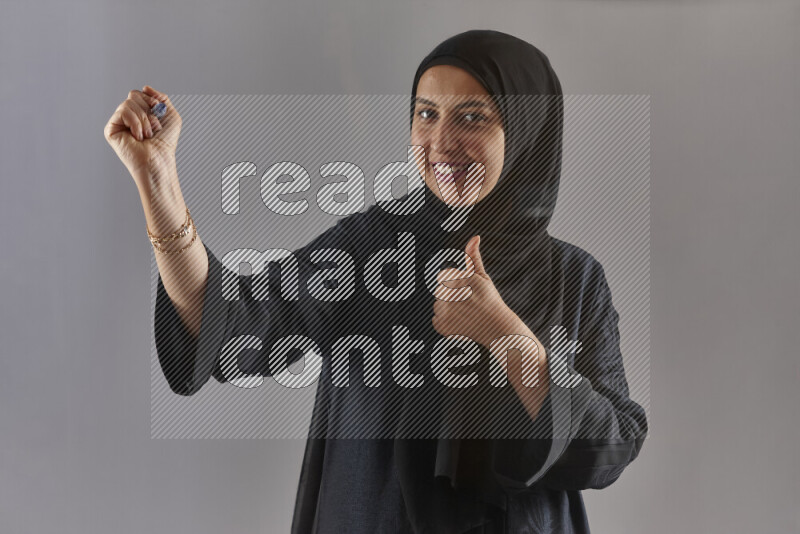 Woman in black abaya and hijab holding a marker pen to write on a transparent board with different reactions and expressions on grey background