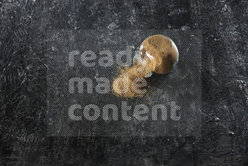 وعاء زجاجي مقلوب ممتلئ ببودرة الكمون والبودرة تناثرت منه على خلفية سوداء