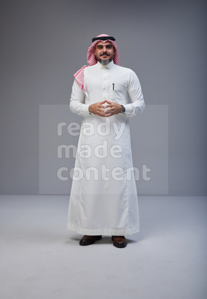 Saudi man Wearing Thob and red white Shomag standing interacting with the camera on Gray background