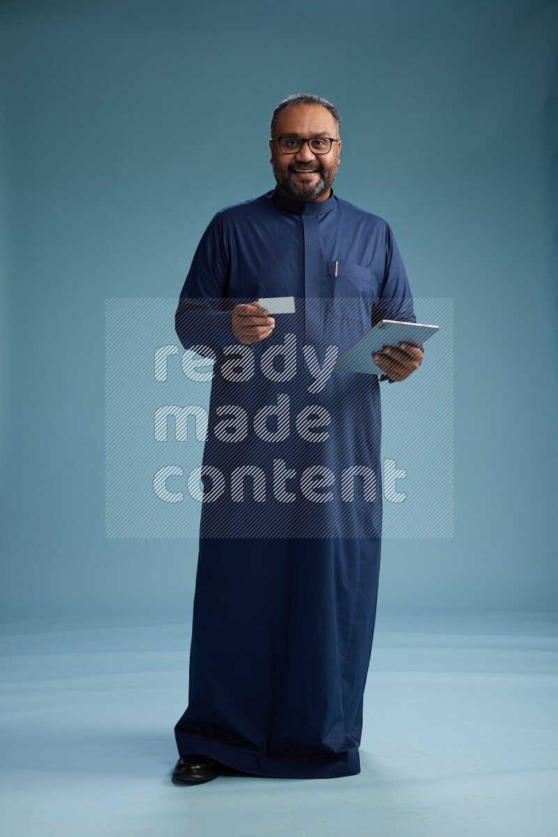 Saudi Man without shimag Standing holding ATM card while working on tablet on blue background