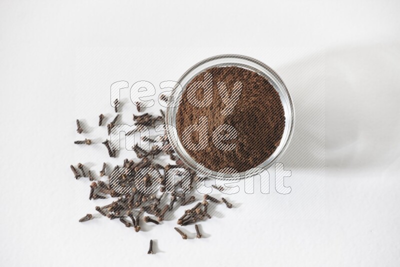 وعاء زجاجي ممتلئ ببودرة القرنفل وحبوب القرنفل متناثرة على أرضية بيضاء