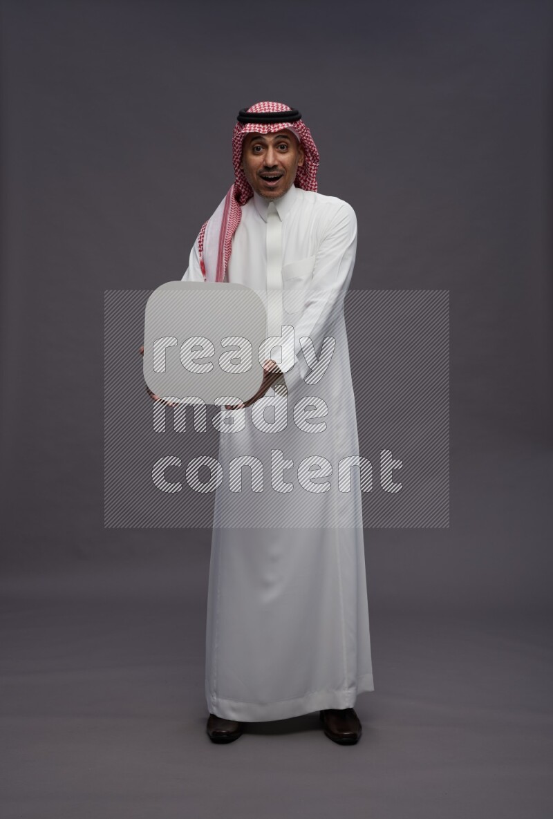 Saudi man wearing thob and shomag standing holding social media sign on gray background