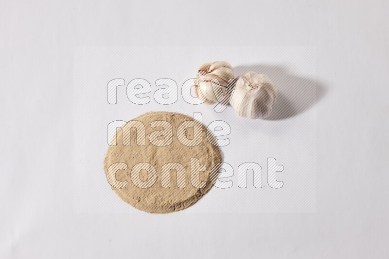 Garlic powder in a circle shape and beside it 2 garlic bulbs on a white flooring