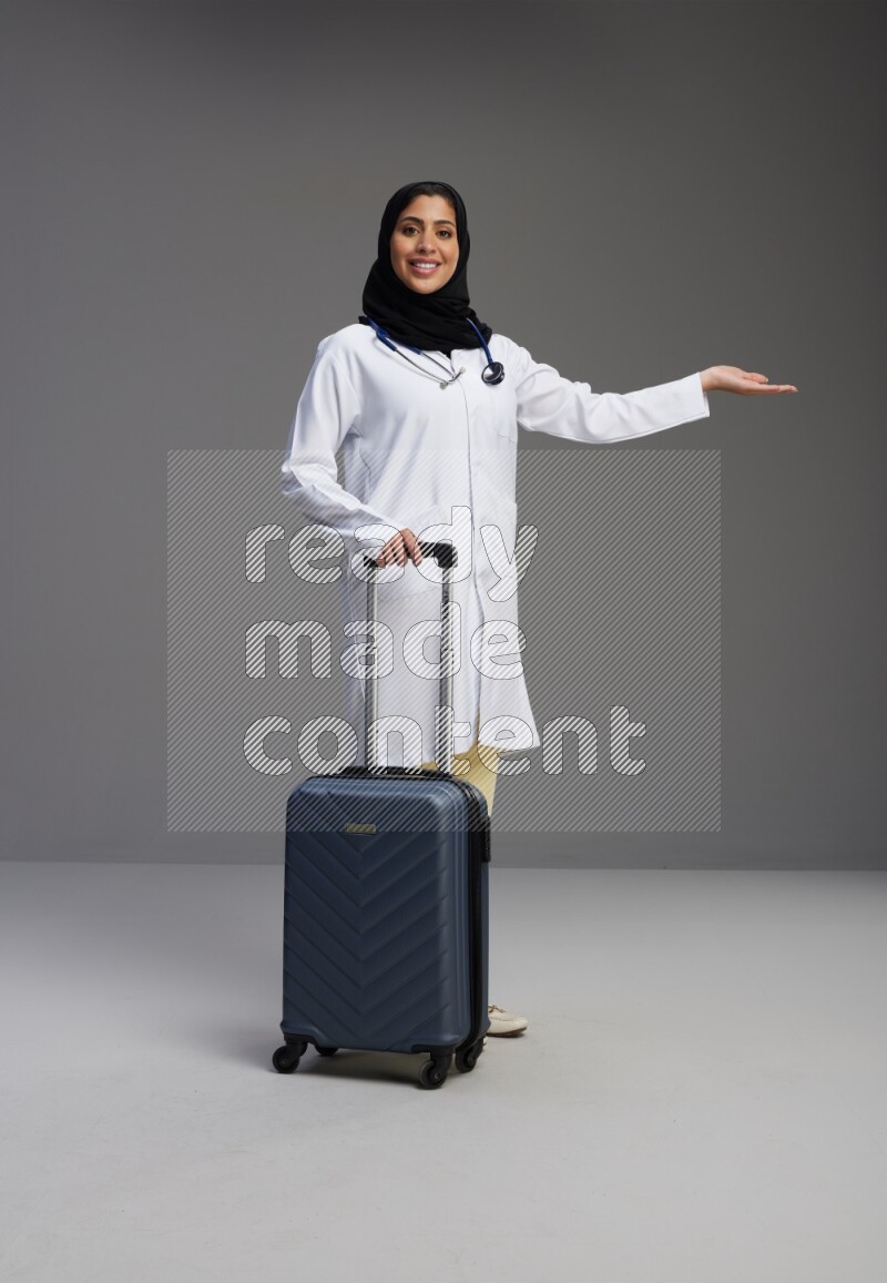 Saudi woman wearing lab coat with stethoscope standing holding Travel bag on Gray background