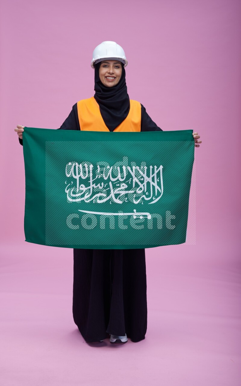 Saudi woman wearing Abaya with engineer vest and helmet standing holding Saudi flag on pink background