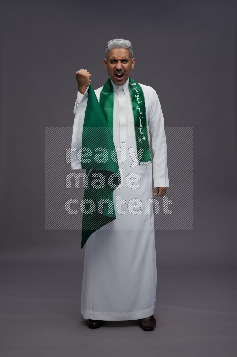 Saudi man wearing thob standing holding Saudi flag on gray background