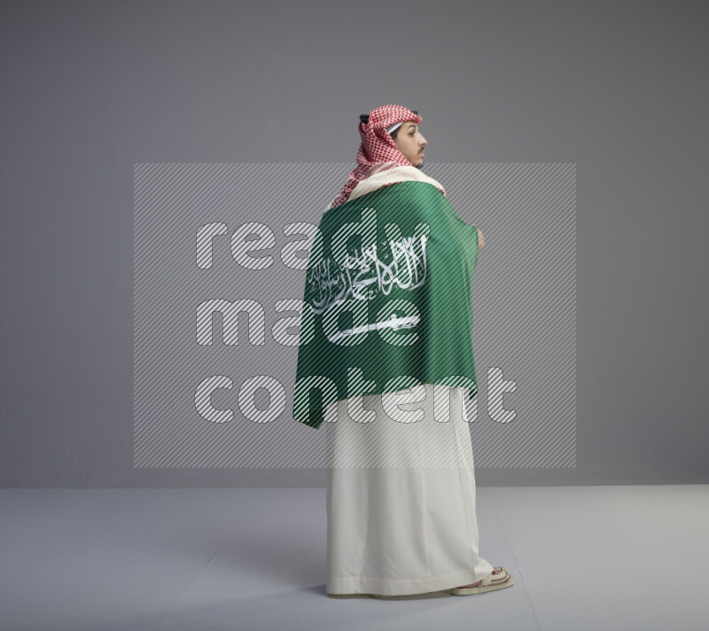 A Saudi man standing wearing thob and red shomag wrapping big saudi flag on gray background