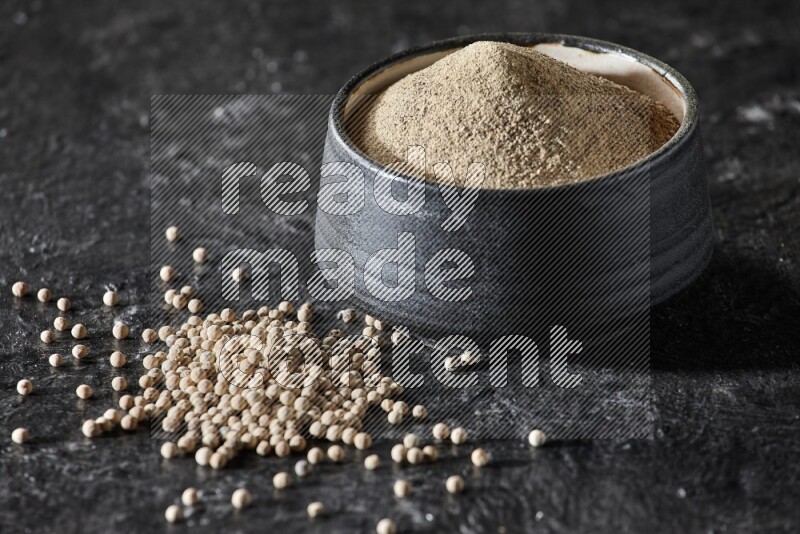 Black pottery bowl full of white pepper powder with white pepper beads on textured black flooring