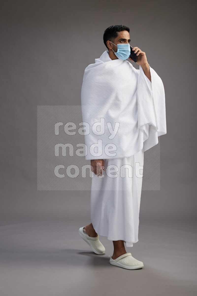 A man wearing Ehram with face mask Standing talking on phone on gray background