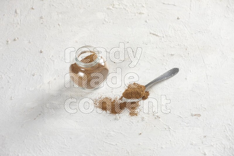 Herbs glass jar full of cinnamon powder with a metal spoon full of powder on a textured white background