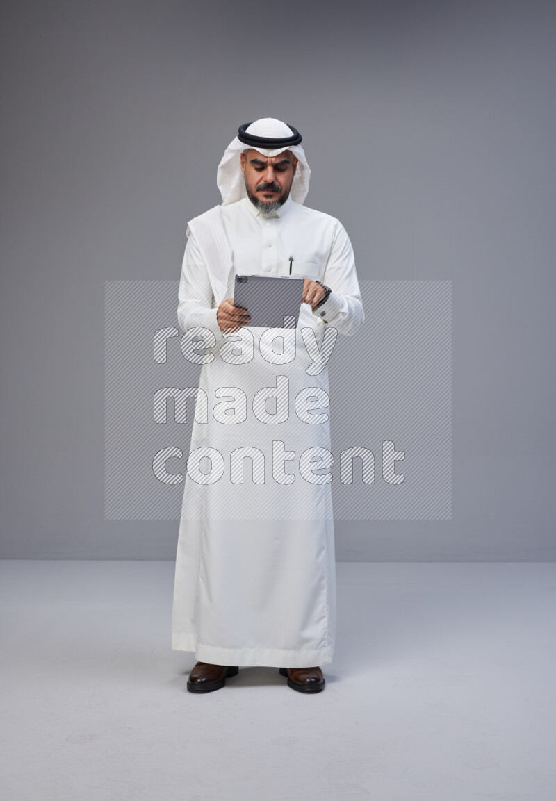 Saudi man Wearing Thob and white Shomag standing working on tablet on Gray background