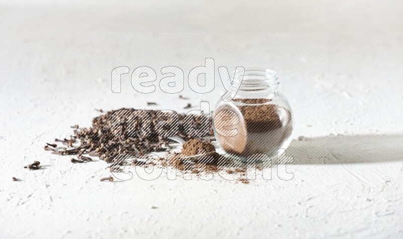 A glass spice jar and a metal spoon full of cloves powder and cloves spread on textured white flooring