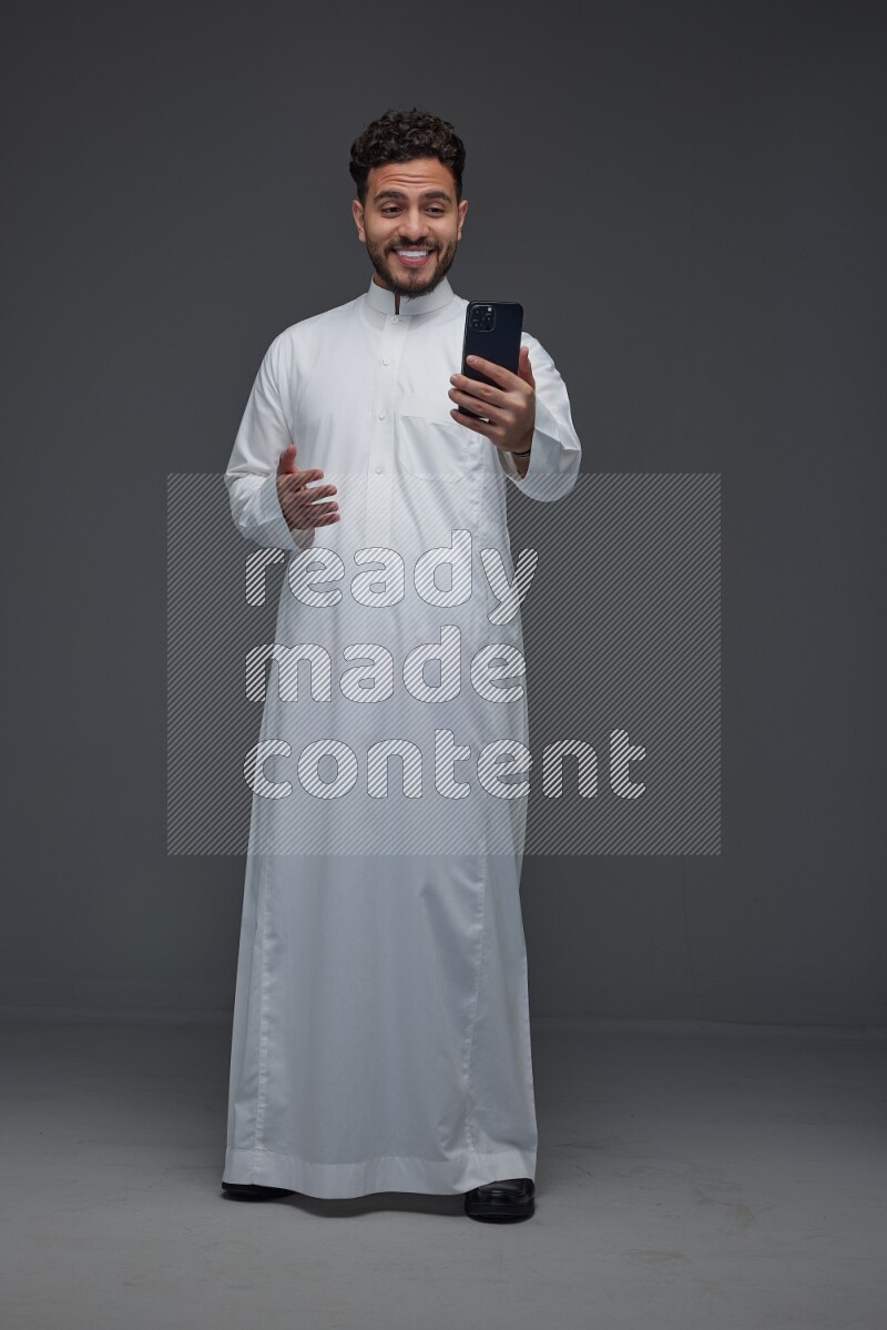 A Saudi man wearing Thobe and making a video call using his phone while standing and making different poses eye level on a gray background