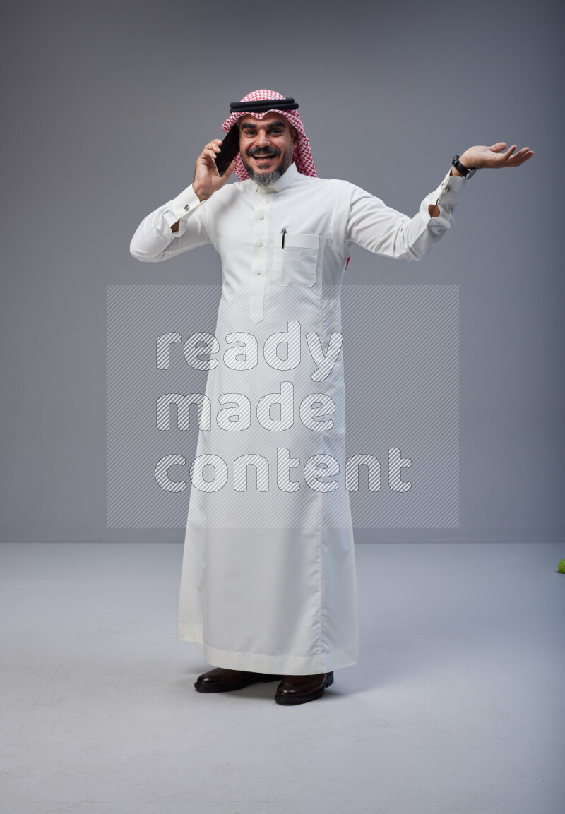 Saudi man Wearing Thob and red Shomag standing talking on phone on Gray background