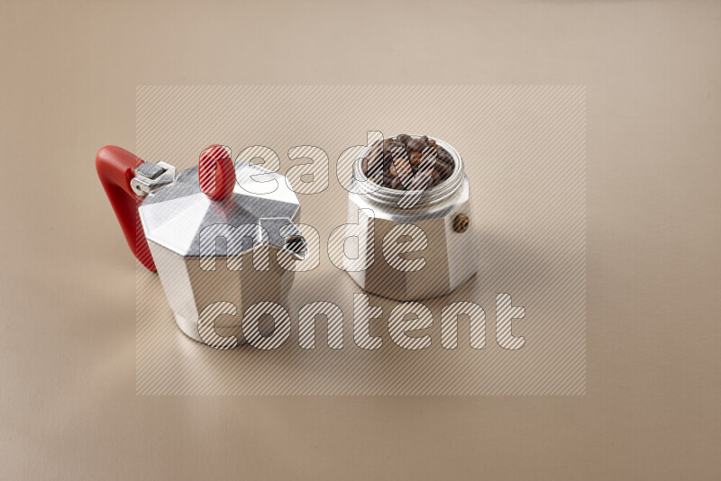 A moka pot with red handle surrounded by roasted coffee beans on beige background