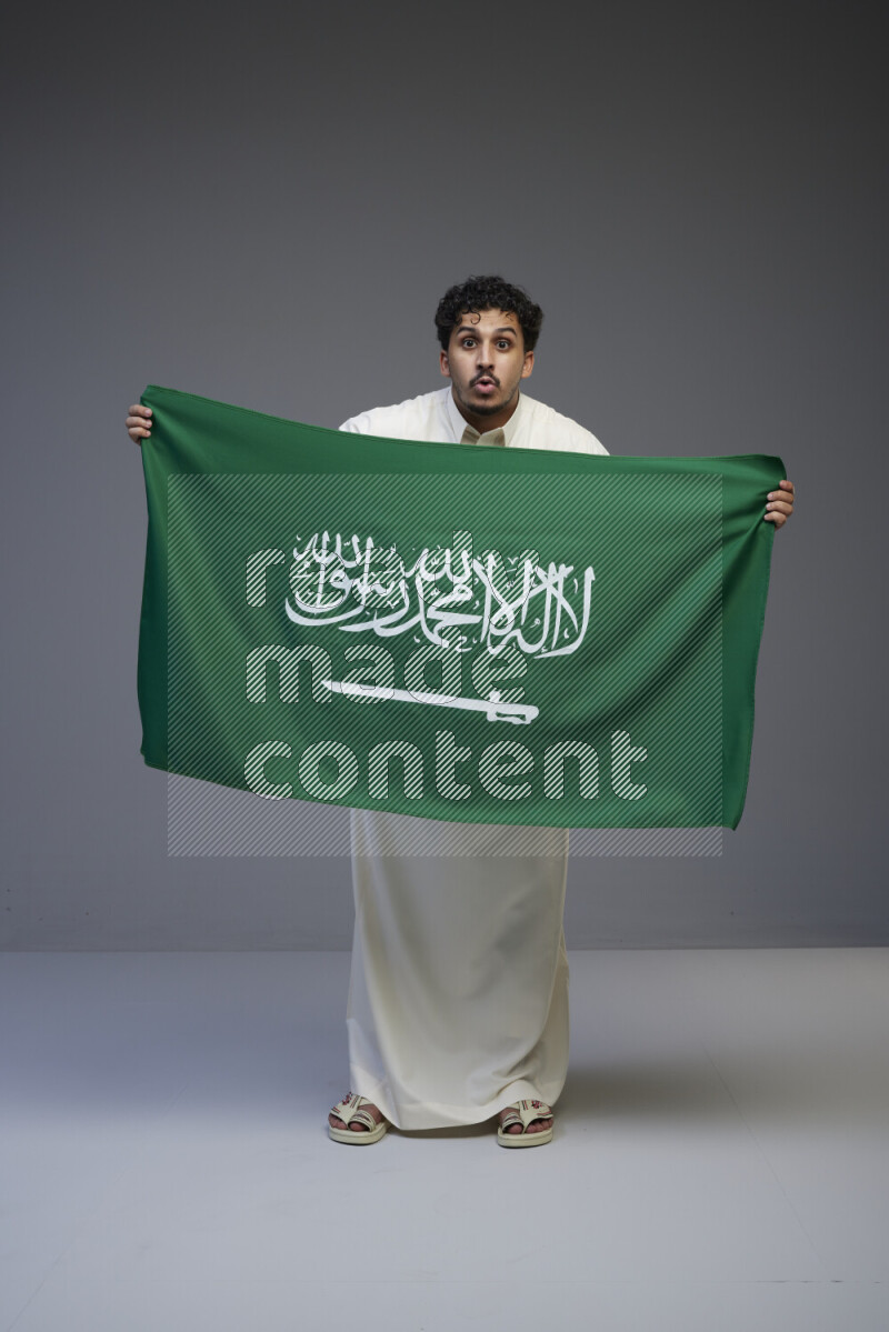 A Saudi man standing wearing thob holding big Saudi flag on gray background