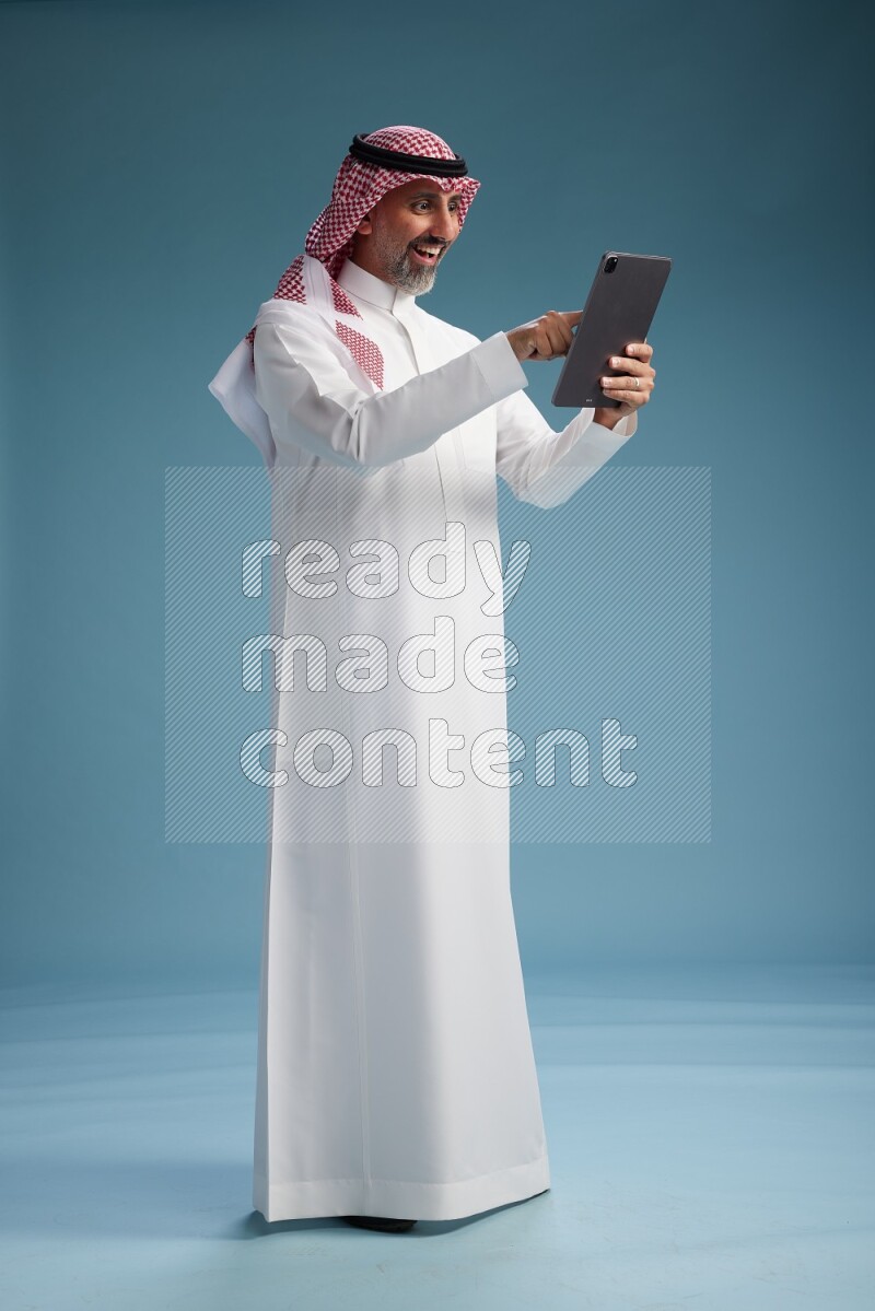 Saudi man wearing thob and shomagh with tablet on Blue background