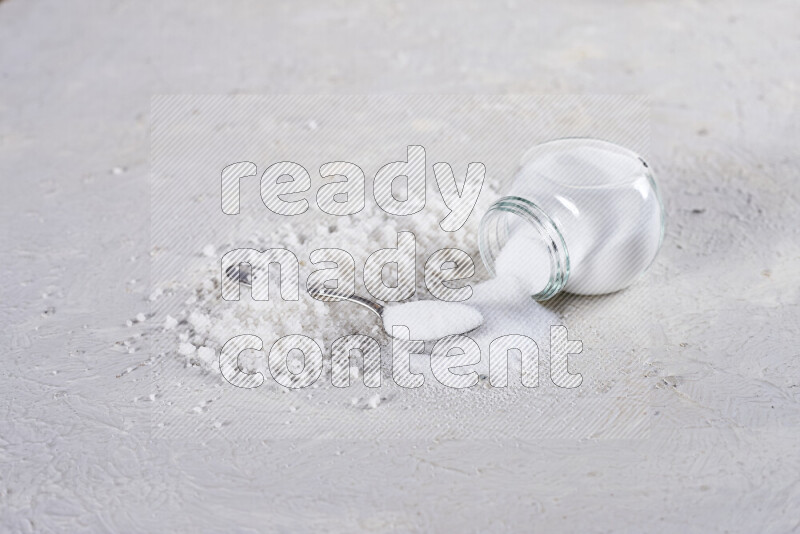 A glass jar full of table salt with some sea salt crystals beside it on a white background